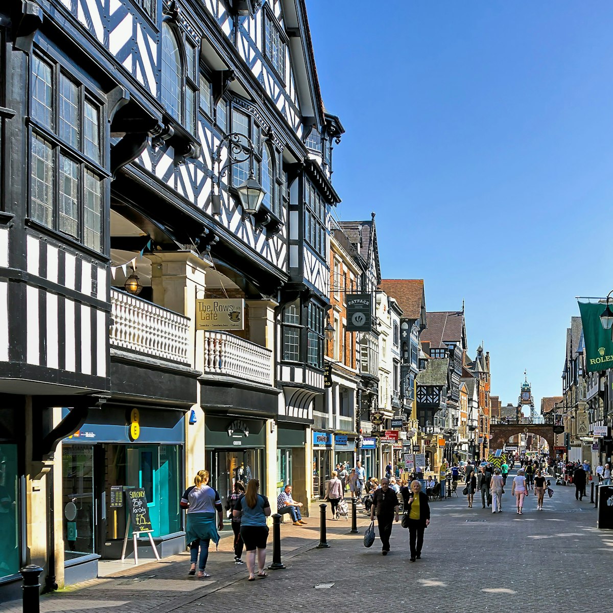 View along the main street in the centre of Chester, Cheshire, UK. Shops can be seen on either side of the road and people can be seen walking and sitting on benches. The rows of upper level shops are pictured on left.
